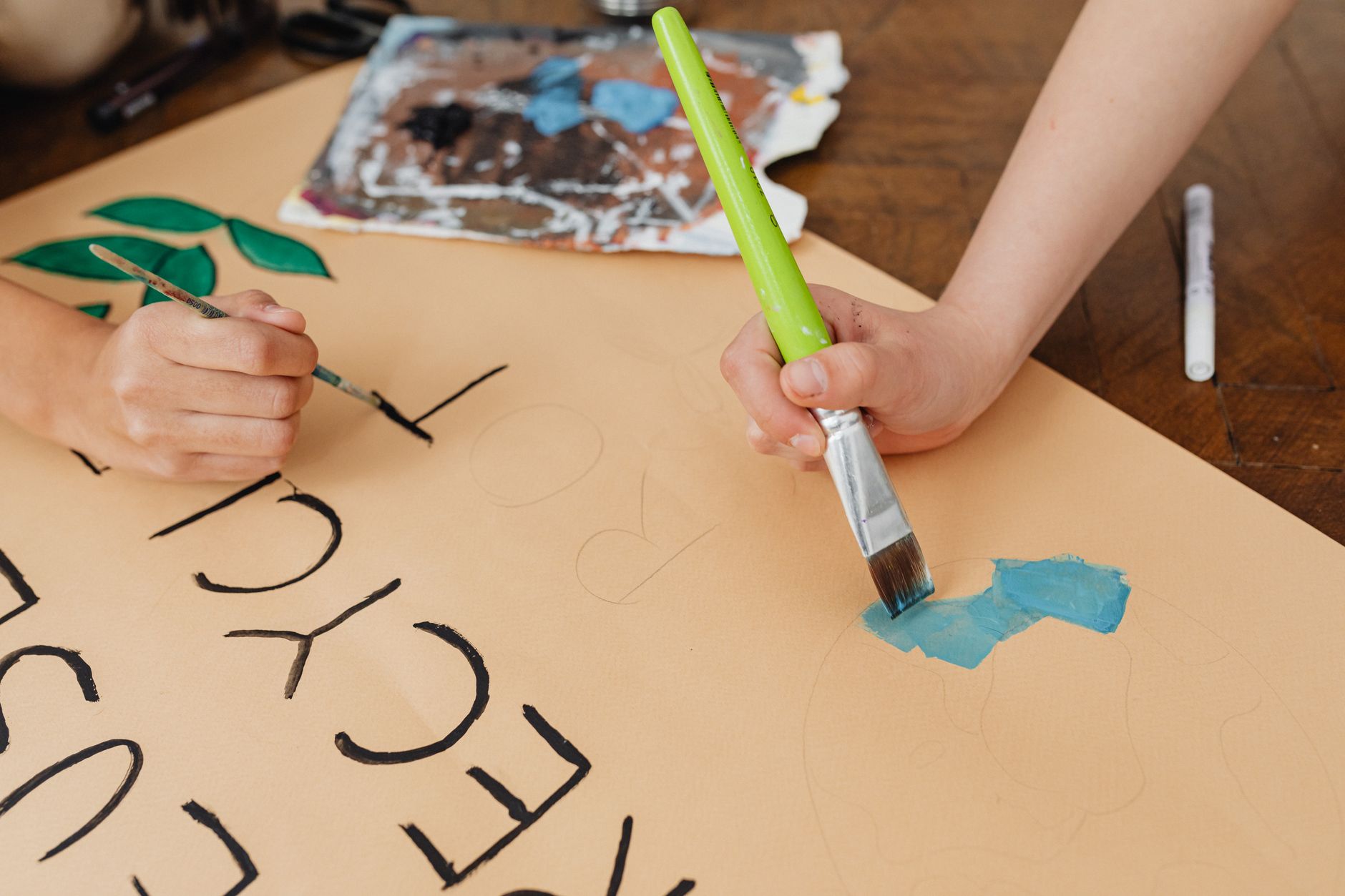 A child's hand painting blue shapes on brown paper with a green-handled brush