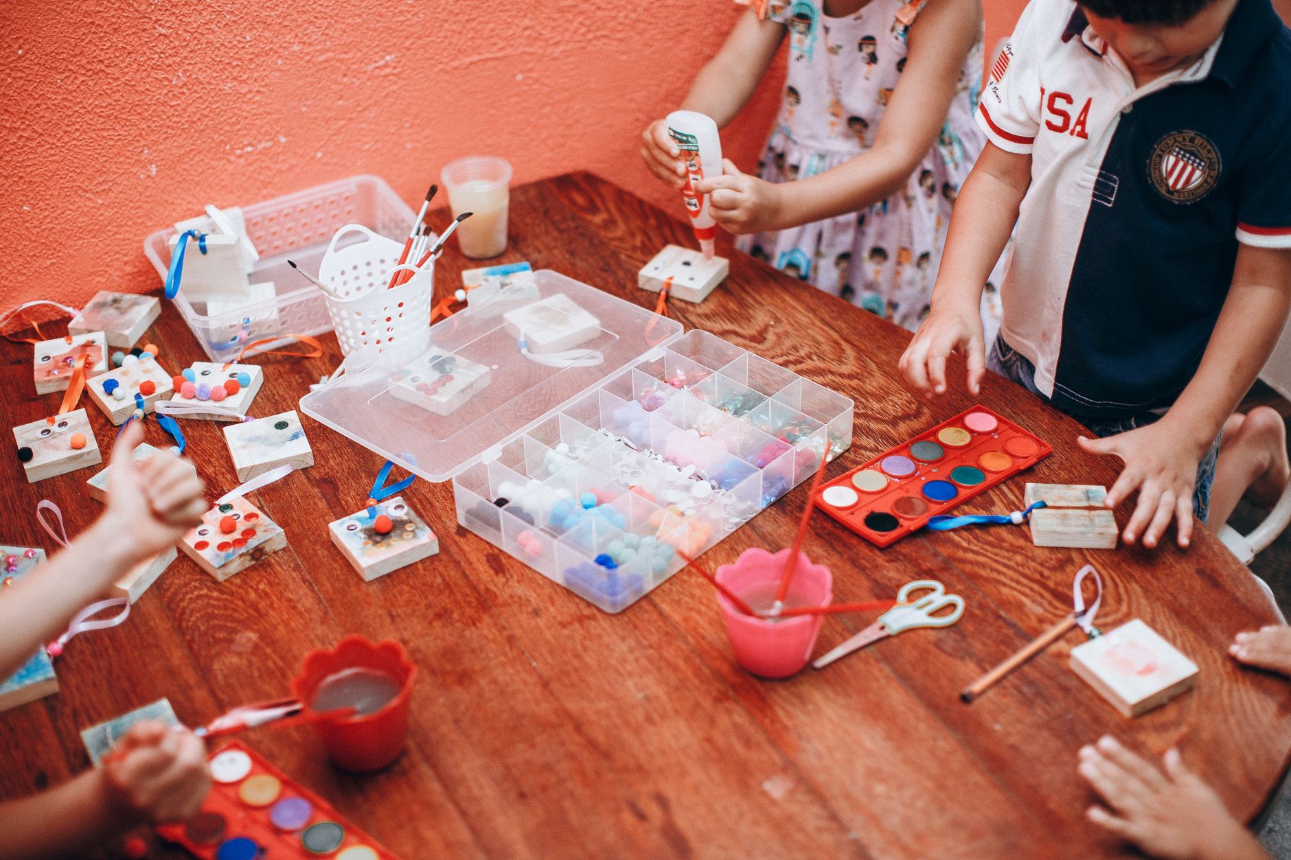 Children gathered around a wooden craft table covered in paint palettes, scissors and art supplies