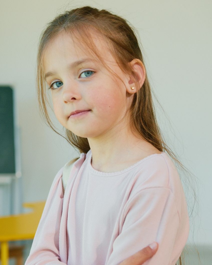 A smiling young girl with long light hair looking at the camera in a bright classroom