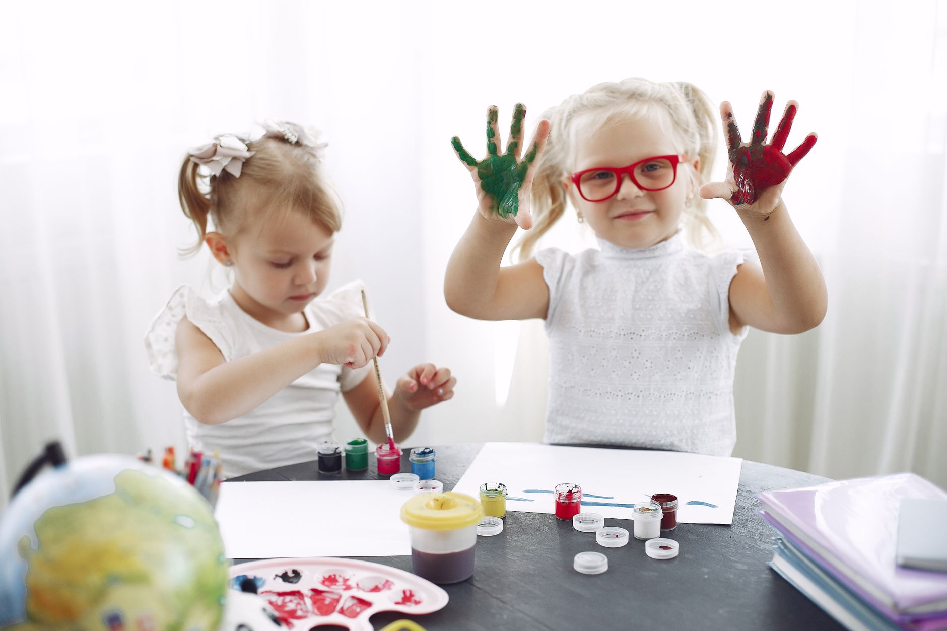 Two little girls holding up hands covered in green and red paint at a children's art table