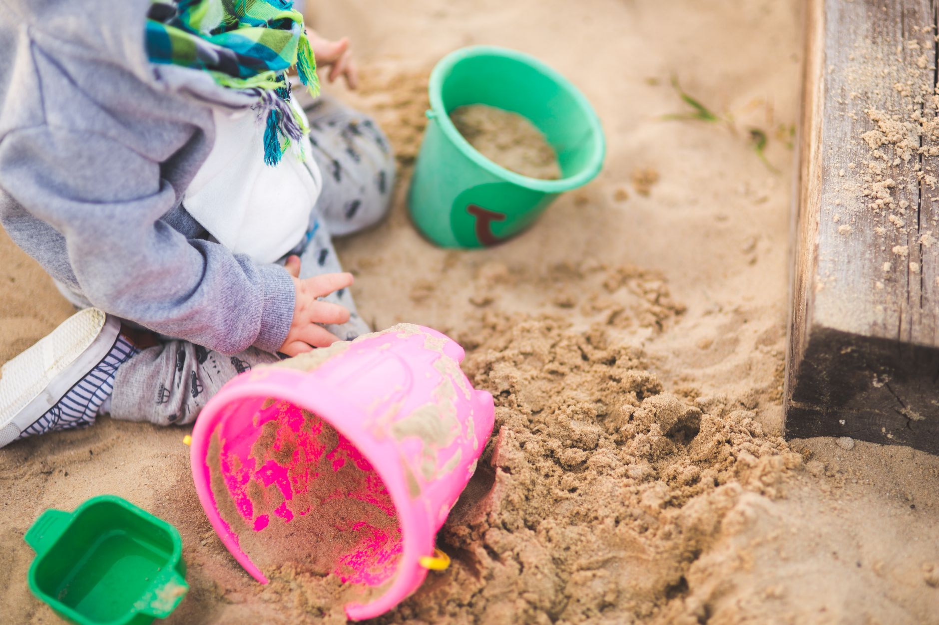 A child playing in a sandpit with a pink bucket and a green bucket