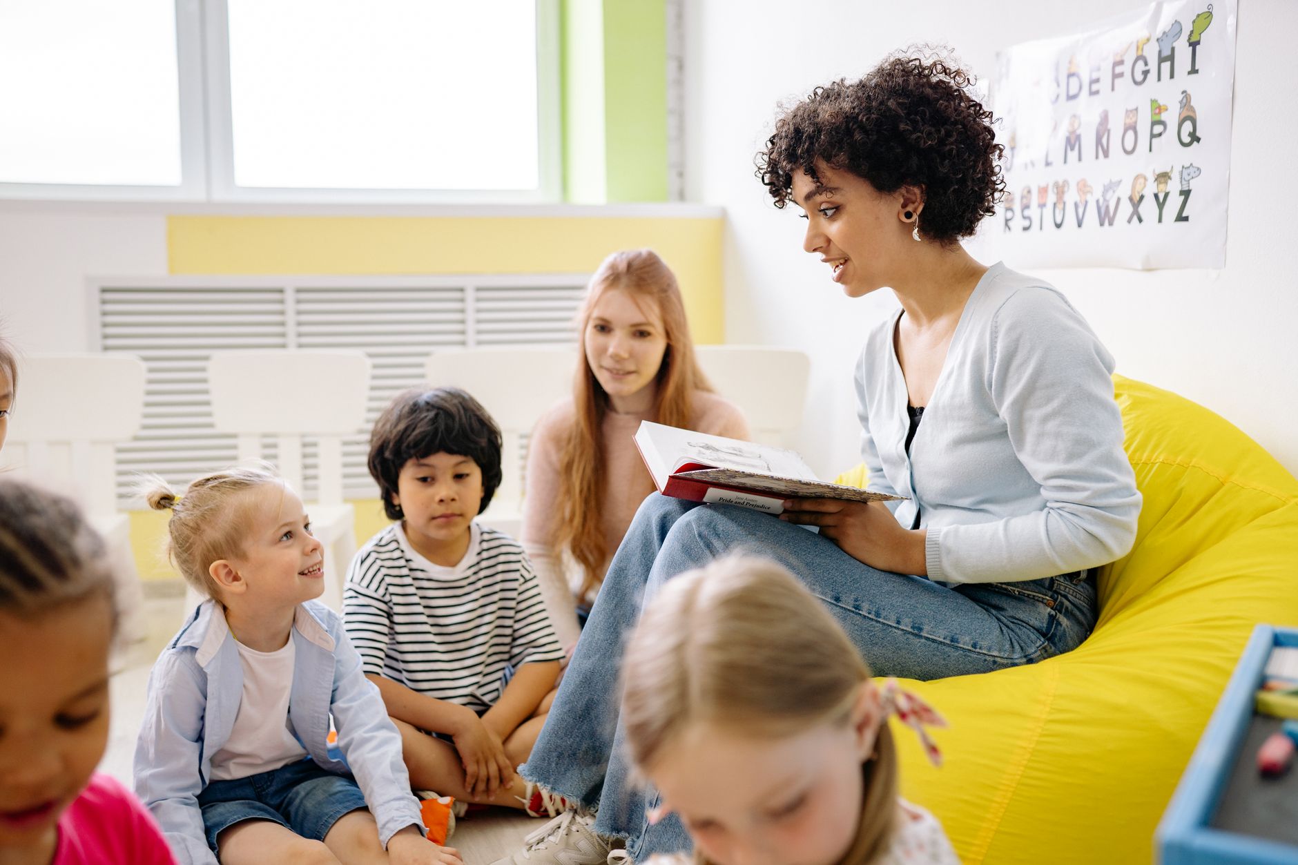 An educator reading to a circle of children on a yellow beanbag in a bright classroom