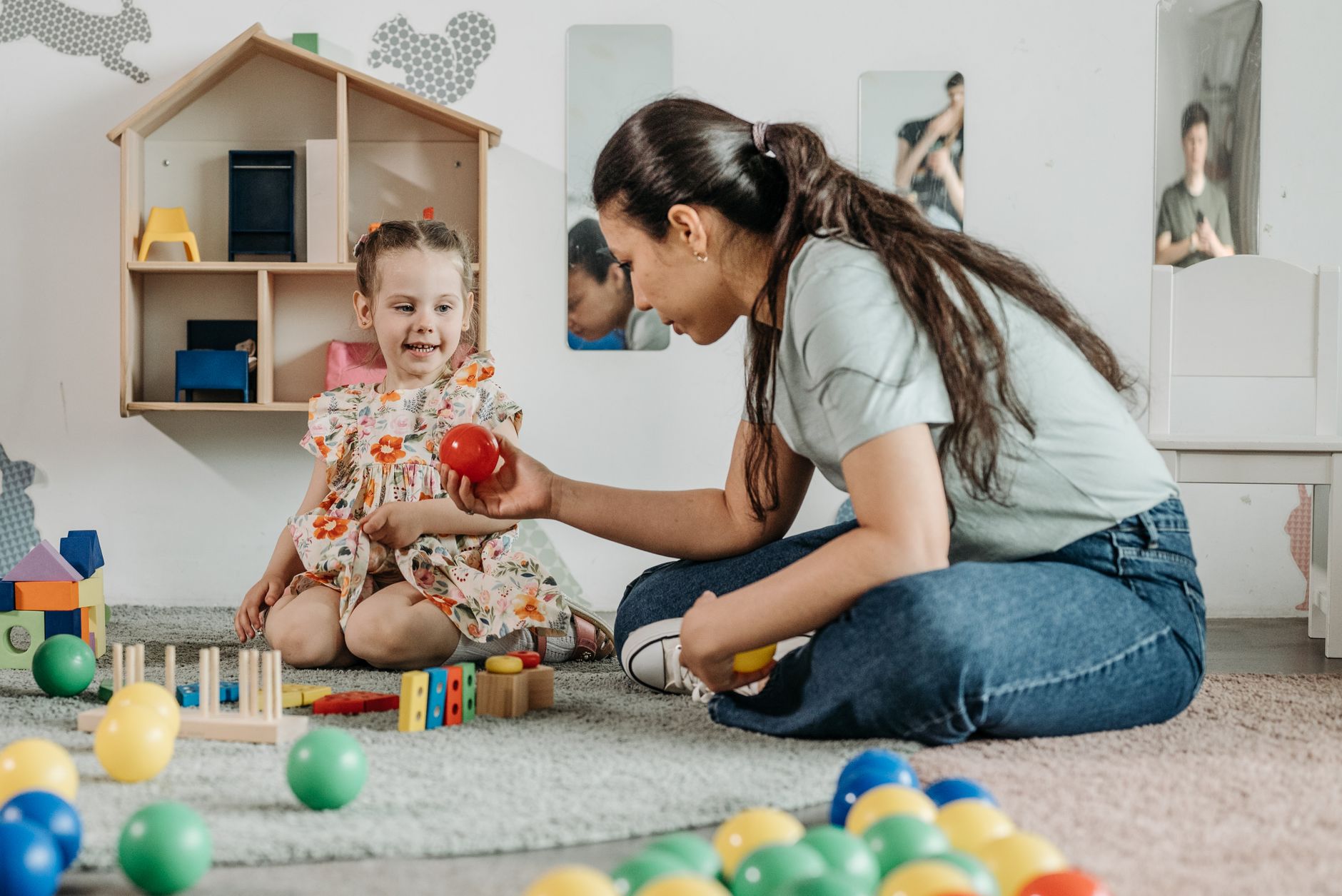 An educator sitting on the floor playing with a little girl and coloured balls